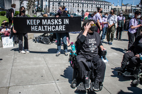 person with black t-shirt that says "Keep Masks in Healthcare in a wheelchair with banner behind